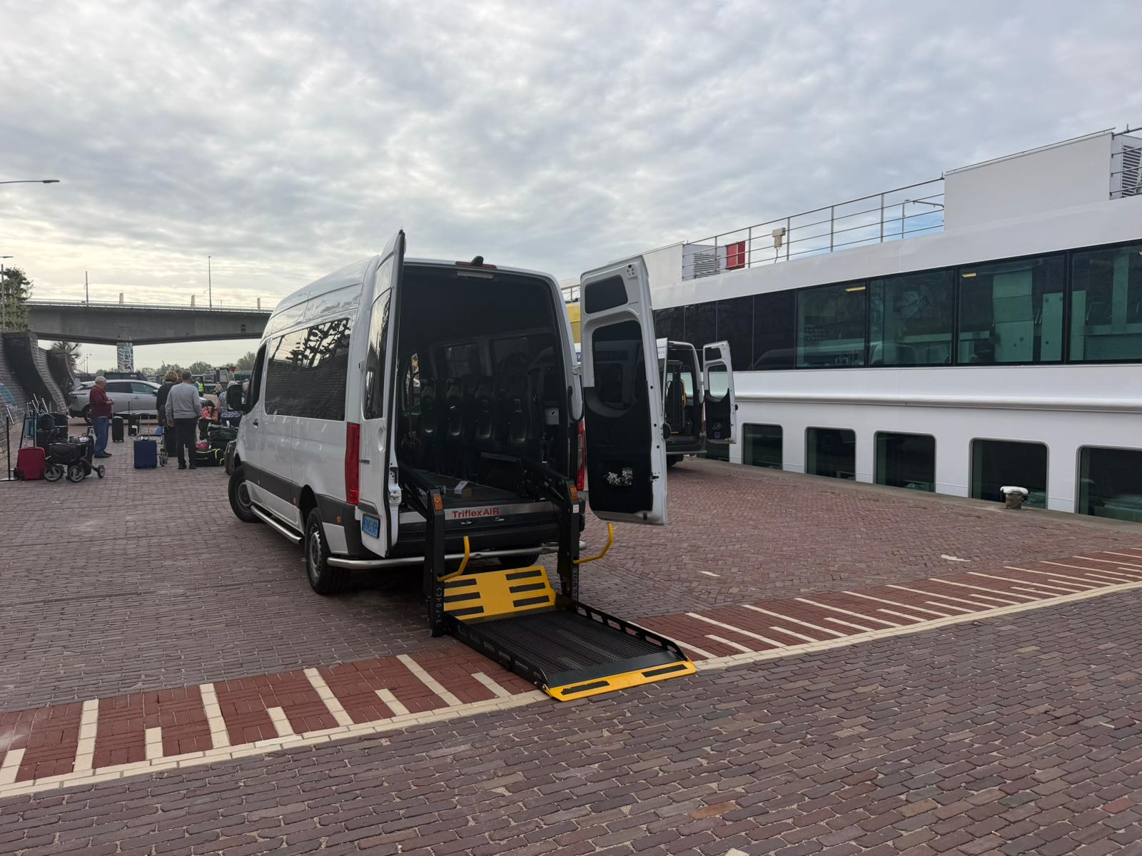 Rolstoelbus met laadklep bij een terminal, reizigers met bagage op de achtergrond
