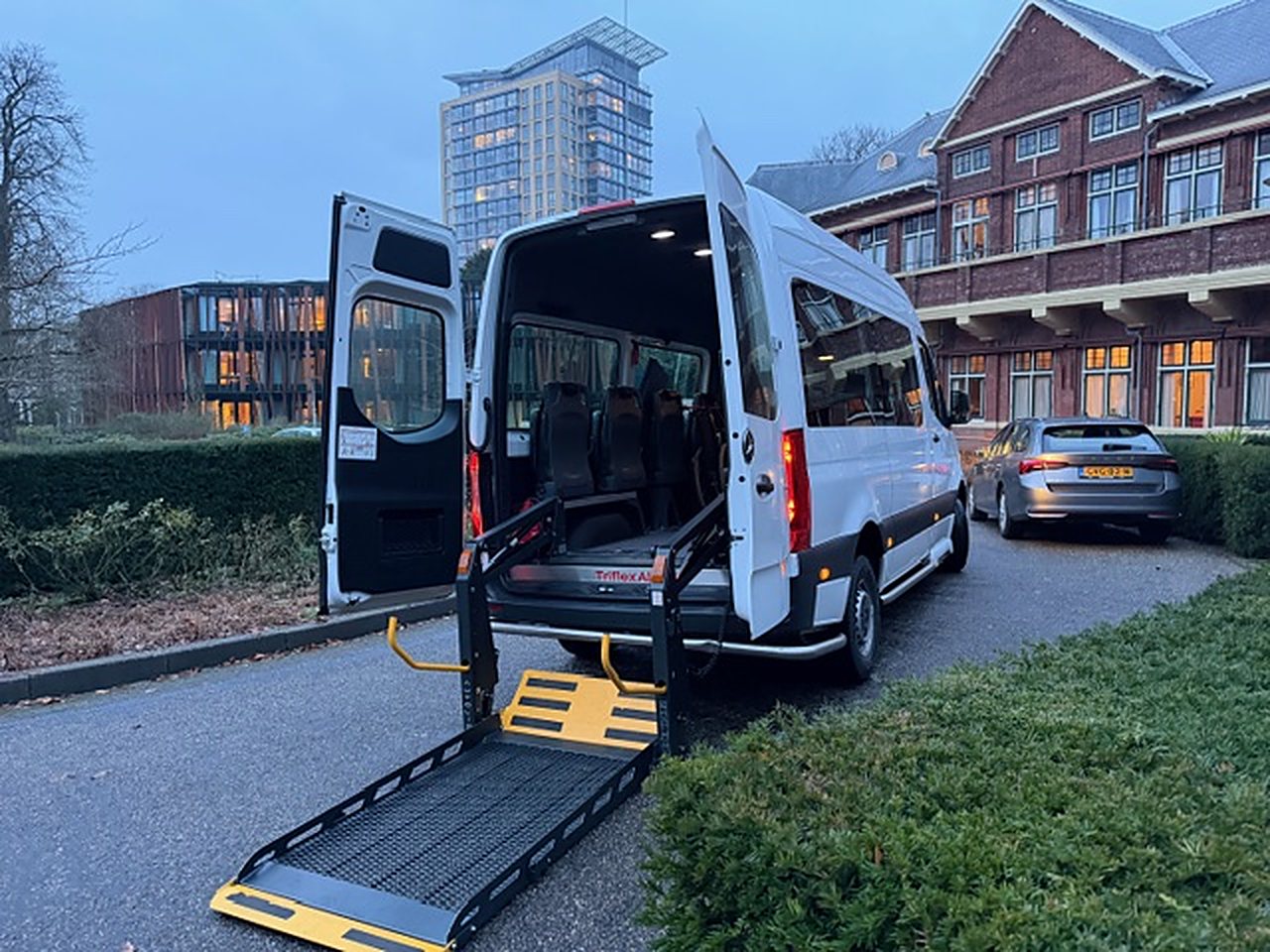 Rolstoelbus met uitgeklapte laadklep, hoogbouw op de achtergrond